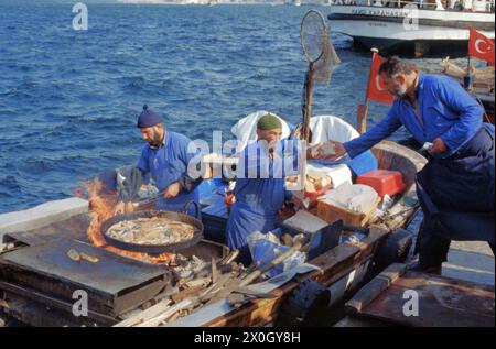 Zwei Männer stehen auf einem Boot am Goldenen Horn in Istanbul in einer großen Pfanne und braten Fisch. Einer der Männer übergibt einem dritten Mann auf dem Steg etwas. Die Männer tragen blaue Overalls. [Automatisierte Übersetzung] Stockfoto