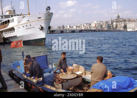 Einige Männer stehen auf einem kleinen Boot am Steg am Goldenen Horn in Istanbul und braten Fisch in einer großen Pfanne. Im Hintergrund die Stadt und der Galata-Turm [automatisierte Übersetzung] Stockfoto