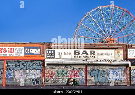 Zwei junge Männer sitzen in der Abendsonne vor einer Reihe geschlossener Restaurants auf dem Coney Island Broadwalk in Brightonbeach, New York. Die Garagentore, die die Geschäfte schließen, sind mit Graffiti bemalt. Im Hintergrund ein Riesenrad mit der Aufschrift „Wonder Wheel“. [Automatisierte Übersetzung]“ Stockfoto