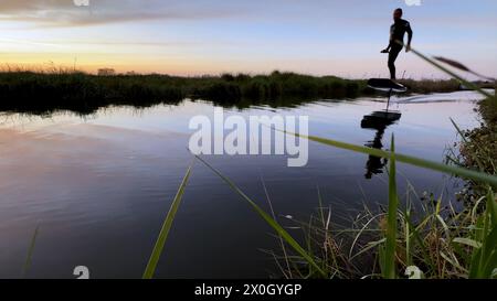 Tragflächenreiter gleitet mit seinem Brett in einem der Kanäle der Ria de Aveiro in Portugal während des Sonnenuntergangs über dem Wasser. Stockfoto