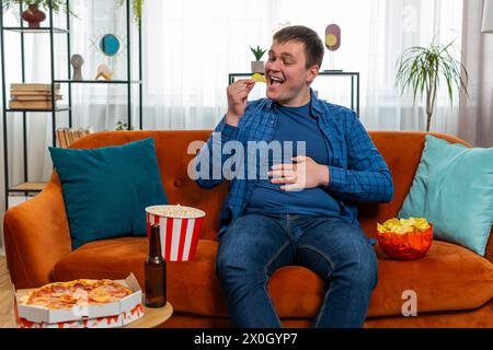 Glücklicher kaukasischer Mann in legeren Klamotten, der auf einem bequemen Sofa sitzt und Chips, Pizza, Popcorn und Bier trinkt, um Filmsport zu sehen und das Wochenende zu Hause zu genießen. Ein lächelnder Kerl isst Fastfood auf der Couch im Haus Stockfoto