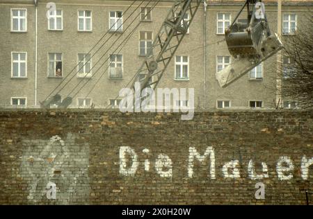 Bau der neuen Berliner Mauer aus Betonfertigteilen. Vor der Mauer befindet sich auf der Ost-Berliner Seite ein Gebäude mit der Aufschrift „die Mauer“, das abgerissen werden soll. Im Hintergrund befindet sich ein Wohngebäude. Stockfoto