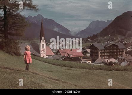 Ein kleines Mädchen im geblümten Kleid blickt auf das Dorf Seefeld in Tyrol mit der Pfarrkirche St. Oswald im Hintergrund. [Automatisierte Übersetzung] Stockfoto