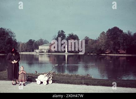 Eine Mutter mit Kind und Kinderwagen stehen am Ufer eines Sees im Park von Schloss Nymphenburg. Im Hintergrund ist ein Teil der Burg. Stockfoto