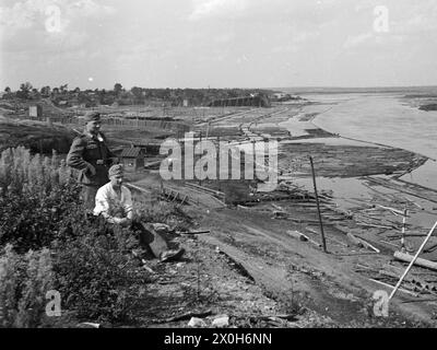 Unzählige Baumstämme werden in einem hölzernen Hafen am Dnieper gesammelt. Zwei deutsche Soldaten sitzen im Gras. Das Bild wurde von einem Mitglied des Radfahrgrenadierregiments 2/Radfahrsicherungsregiment 2 im Nordteil der Ostfront aufgenommen. [Automatisierte Übersetzung] Stockfoto