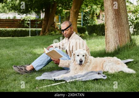 Ein Afroamerikaner mit Myasthenia gravis-Syndrom sitzt im Gras mit seinem Labrador, vertieft in ein Buch. Stockfoto