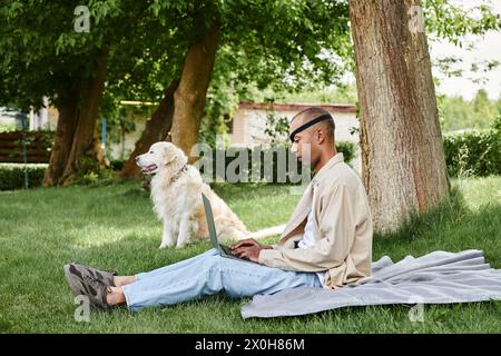 Ein vielseitiger Mann mit Myasthenia gravis sitzt auf dem Gras und benutzt einen Laptop, während er von seinem treuen Labrador-Hund begleitet wird. Stockfoto