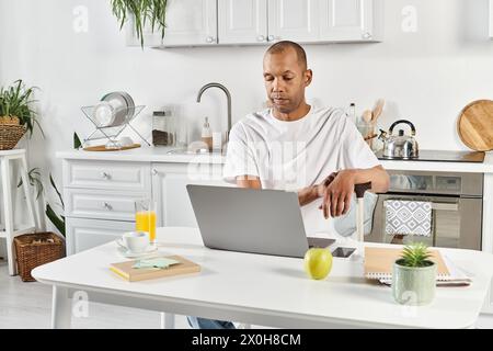 Ein vielseitiger Afroamerikaner mit Myasthenia gravis sitzt an einem Küchentisch, vertieft in seinen Laptop. Stockfoto