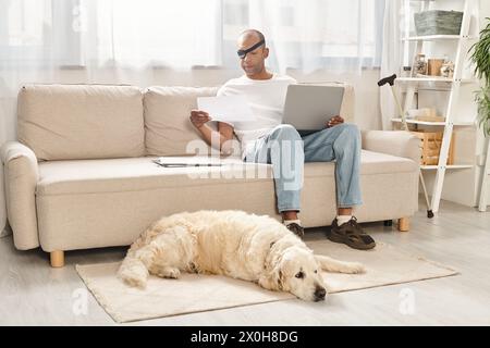 Ein Mann, der gegen Myasthenia gravis kämpft, sitzt auf einer Couch mit einem Laptop, begleitet von seinem treuen Labrador-Hund. Stockfoto