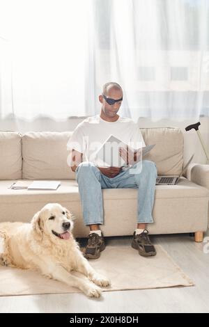 Ein behinderter afroamerikanischer Mann mit Myasthenia gravis-Syndrom, der auf einer Couch neben einem Labrador-Hund sitzt und Vielfalt und Inklusion verkörpert. Stockfoto