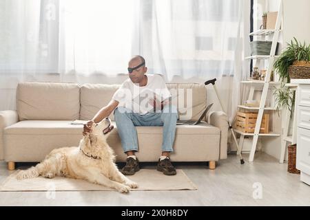 Ein behinderter Afroamerikaner mit Myasthenia gravis-Syndrom sitzt auf einer Couch neben seinem Labrador-Hund. Stockfoto