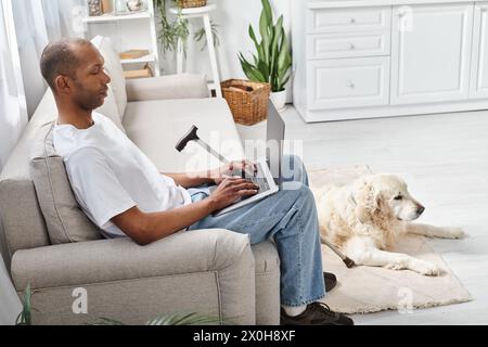 Ein Afroamerikaner mit Myasthenia gravis, der auf einer Couch sitzt und einen Laptop neben seinem Labrador-Hund benutzt. Stockfoto