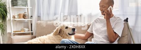 Ein Mann mit Myasthenia gravis sitzt auf einer Couch und spricht auf einem Handy neben seinem loyalen Labrador-Hund. Stockfoto