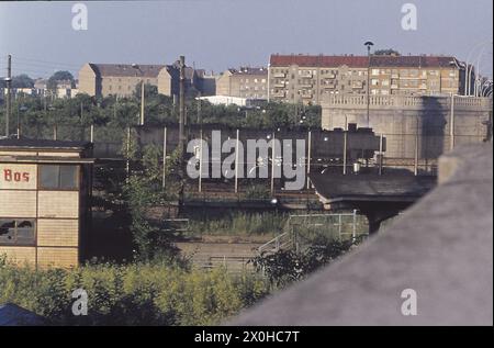 Das Bild wurde 1977 von West-Berlin in Richtung Ost-Berlin am Bahnhof Bornholmerstraße aufgenommen, dessen Bahnsteig und baufälliges Stellwerk gerade zwischen dem Grün erkennbar sind. Da die Grenze mitten durch den großen Streckenverteiler zur Nordbahn verläuft, verlagerte die DR eine Reihe von Gleisen auf Ost-Berliner Territorium, so dass die Züge nicht durch West-Berlin fahren mussten. Der Grenzzaun verläuft entlang der eigentlichen Grenzlinie. Die Mauer selbst ist im Hinterland unsichtbar. [Automatisierte Übersetzung] Stockfoto