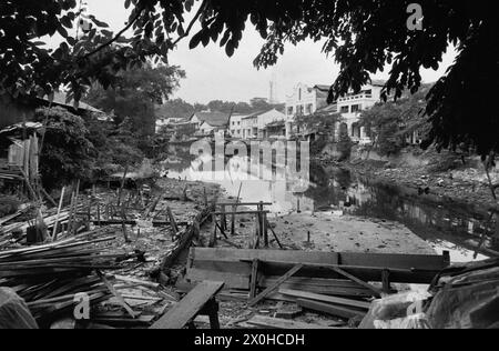 Die Überreste von Holzhütten am Wasser in Singapur. Nur einzelne Holzbretter und Pfosten sind noch sichtbar. Im Hintergrund intakte Steinhäuser. [Automatisierte Übersetzung] Stockfoto