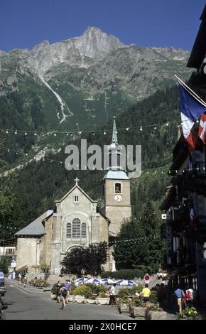 Die Pfarrkirche von Chamonix mit Le BrÃ Vent im Hintergrund. Menschen auf der Straße, Tische und weiß-blaue Sonnenschirme vor dem Hotel Le Chamonix. [Automatisierte Übersetzung] Stockfoto