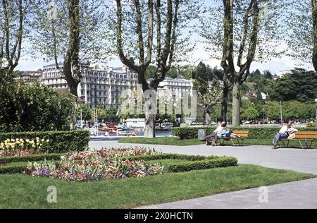 An der Seepromenade in Ouchy. Blick auf den Genfer See und das Hotel Beau-Rivage Palace von einem grünen Bereich mit Blumenbeeten. Ein Mann und eine Frau schlafen auf einer Parkbank, ein Mann sitzt auf der anderen Bank [automatisierte Übersetzung] Stockfoto