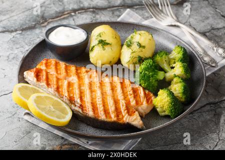 Gegrillter Lachs mit gekochten Kartoffeln, Brokkoli, Zitrone, Kräutern und Sahnesauce in Nahaufnahme auf einem Teller auf dem Tisch. Horizontal Stockfoto