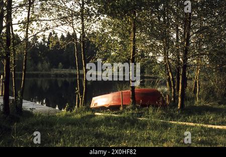 Ein umgedrehtes rotes Ruderboot liegt am Rande eines kleinen, idyllischen Sees [automatisierte Übersetzung] Stockfoto