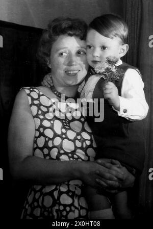 Eine Mutter in Ochsenfurt trägt ihren Sohn in den Armen, während er Blumen hält. [Automatisierte Übersetzung] Stockfoto