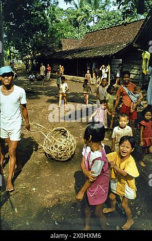 Eine Gruppe von Kindern und Jugendlichen lachen über einen Platz in einem Dorf in der Nähe von Yogyakarta in Indonesien, EIN junger Mann zieht einen gewebten Korb hinter sich. Die Holzhäuser stehen auf einer Lichtung im Wald. [Automatisierte Übersetzung] Stockfoto