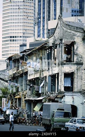 Blick auf eine Straße in Singapur. An der Straße befinden sich mehrere alte und heruntergekommene Mietshäuser. Die Holzläden sind geöffnet. Wäsche hängt an langen Stangen aus den Fenstern, um zu trocknen. Fußgänger und Fahrzeuge sind unterwegs. Im Hintergrund sind Fassaden moderner Hochhäuser zu sehen. [Automatisierte Übersetzung] Stockfoto