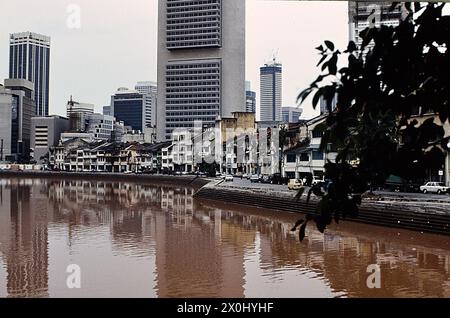 Blick entlang einer Strandpromenade in Singapur. Im Vordergrund rotbraunes Wasser. An der Bank eine Straße. Dahinter alte Häuser mit heruntergekommenen Fassaden und Dächern. Im Hintergrund neue Bürohochhäuser. [Automatisierte Übersetzung] Stockfoto