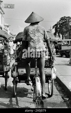 Ein Mann reitet auf einer weit entfernten Rikscha durch die Straßen von Yogyakarta. Fahrer und Rad sind nur von hinten im Bild zu sehen. Der Mann trägt ein gemustertes Hemd, eine kurze Hose und einen Reishut. [Automatisierte Übersetzung] Stockfoto