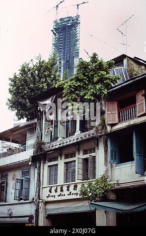 Ein altes Apartmentgebäude in Singapur. Auf dem Haus wachsen Sträucher und kleine Bäume. Im Hintergrund sieht man den Neubau eines modernen Hochhauses. Oben auf dem Wolkenkratzer befinden sich zwei Baukräne. [Automatisierte Übersetzung] Stockfoto