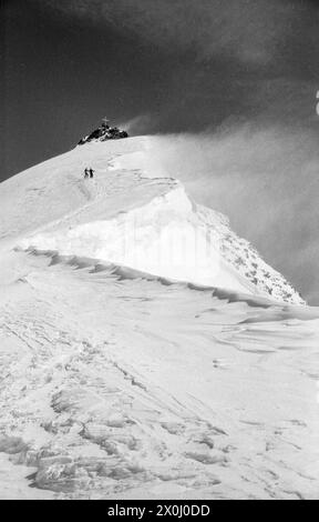 Zwei Skitourenfahrer steigen von der Wildspitze ab. Der Gipfel mit Gipfelkreuz im Hintergrund. Der Wind weht Schnee vom Kamm. [Automatisierte Übersetzung] Stockfoto