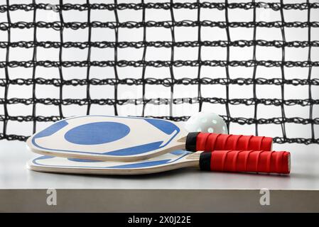 Zwei blaue und rote Pickleball-Schläger und ein weißer Ball auf weißem Tisch, isoliert mit einem Spielnetzwerk im Hintergrund. Vorderansicht. Stockfoto