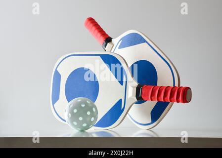 Zwei blaue und rote Pickleball-Schläger und eine weiße Kugel auf weißem Tisch und weißem isoliertem Hintergrund. Vorderansicht. Stockfoto