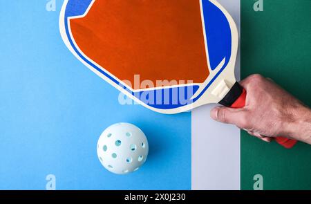 Hintergrund mit Hand mit Pickleball-Paddel auf grüner und blauer Spielfläche mit weißem Ball. Draufsicht. Stockfoto