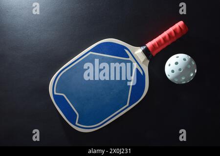 Blauer und roter Holzpaddel-Pickleball-Schläger und eine weiße Kugel auf schwarzem Tisch. Draufsicht. Stockfoto