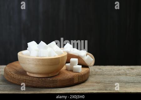 Weiße Zuckerwürfel in Schüssel und Schaufel auf Holztisch vor dunklem Hintergrund, Nahaufnahme. Leerzeichen für Text Stockfoto