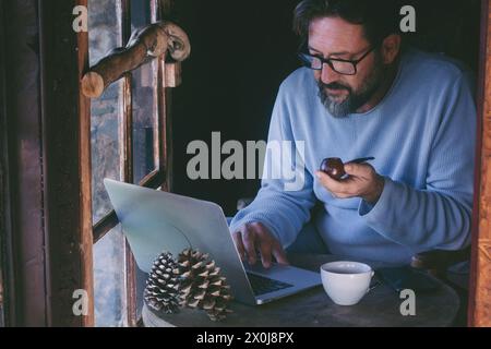 Ein reifer Mann sitzt zu Hause und arbeitet an einem Laptop-Computer und raucht Pfeife. Alternative Single Lifestyle männliche Leute innen in gemütlichem Hütte Holzchalet. Telearbeit online Stockfoto