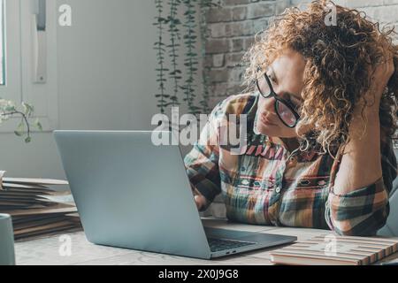 Eine moderne freischaffende Unternehmerin, die zu Hause an einem Laptop arbeitet, in einem kleinen Büro, berührt ihren Kopf und schaut mit Zweifeln und Neugier auf den Display-Computer. Online-Frauen Stockfoto