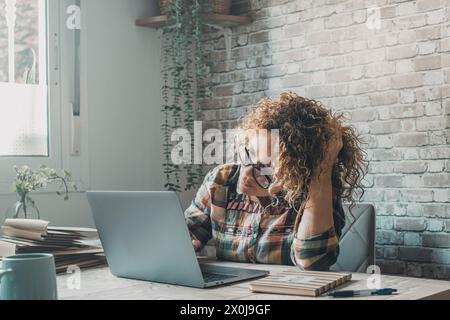 Eine moderne freischaffende Unternehmerin, die zu Hause an einem Laptop arbeitet, in einem kleinen Büro, berührt ihren Kopf und schaut mit Zweifeln und Neugier auf den Display-Computer. Online-Frauen Stockfoto