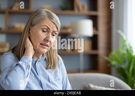 Eine ältere kaukasische Frau, die in einem Wohnzimmer sitzt, scheint beunruhigt und unwohl zu sein und hat möglicherweise Nackenschmerzen oder allgemeines Unwohlsein. Stockfoto