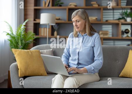 Eine reife Frau, konzentriert und engagiert, benutzt einen Laptop, während sie in einem gut eingerichteten Wohnzimmer auf einem Sofa sitzt, was eine Mischung aus Komfort und Produktivität andeutet. Stockfoto