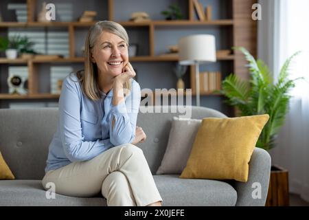 Eine fröhliche Seniorin sitzt bequem auf einem grauen Sofa zu Hause, sieht besinnlich und lächelnd aus, in einem gut dekorierten Wohnzimmer mit modernem Flair. Stockfoto