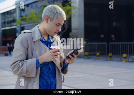 Junger blonder lateinamerikanischer Universitätsmann, in formellen Kleidern, stehend und recherchiert sein Handy vor der Universität, surft im Internet, sucht soziale Netzwerke, kopiert Stockfoto