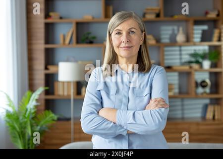 Porträt einer selbstbewussten älteren Frau mit überkreuzten Armen in einem stilvollen, modernen Wohnzimmer. Ihr heiterer Ausdruck strahlt Professionalität und Erfahrung aus. Stockfoto