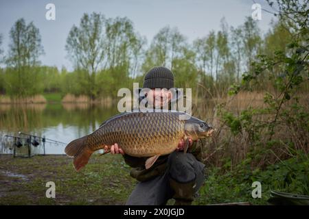 Fischer, der einen Karpfen mit einer Seenlandschaft im Hintergrund hält. Modellversion verfügbar. Stockfoto