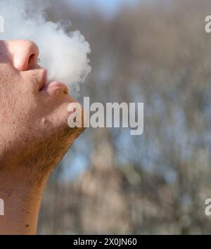 Nahaufnahme eines jungen Mannes, der eine elektronische Zigarette raucht. Junger Mann im Profil, der das Ausatmen von dampfendem Rauch genießt. Stockfoto