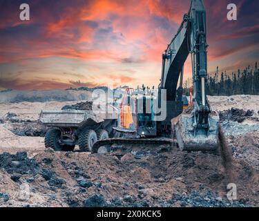 Bagger gräbt bei Sonnenuntergang Erde auf einer Baustelle, während ein Kipplaster im Hintergrund vorbeifährt Stockfoto