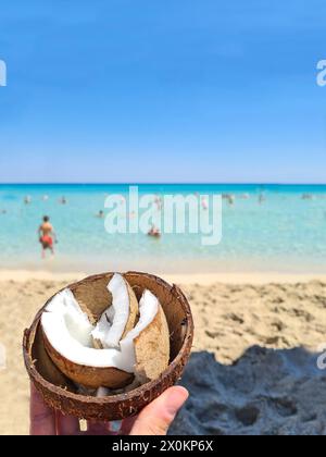 Eine Frauenhand hält eine offene Kokosnuss mit Muscheln im Vordergrund, ein typischer Snack am Strand während des Sommerurlaubs auf Mallorca, Spanien Stockfoto