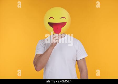 Mann, der das Gesicht mit Emoticon bedeckt, der die Zunge auf gelbem Hintergrund hinausragt Stockfoto