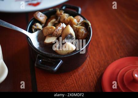 Köstliche Pilzscheiben in einem schwarzen Topf gekocht, heiß mit einem Löffel auf einem Holztisch serviert. Vegetarische Beilagen im argentinischen Steakhouse Restaurant Stockfoto