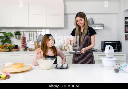 Zwei junge Frauen machen Nachtisch in der Küche. Eine Person, die einen weißen Kuchenteig oder eine Glasmischung in einer klaren Glasschale umrührt. Der andere Teig wird gegossen Stockfoto